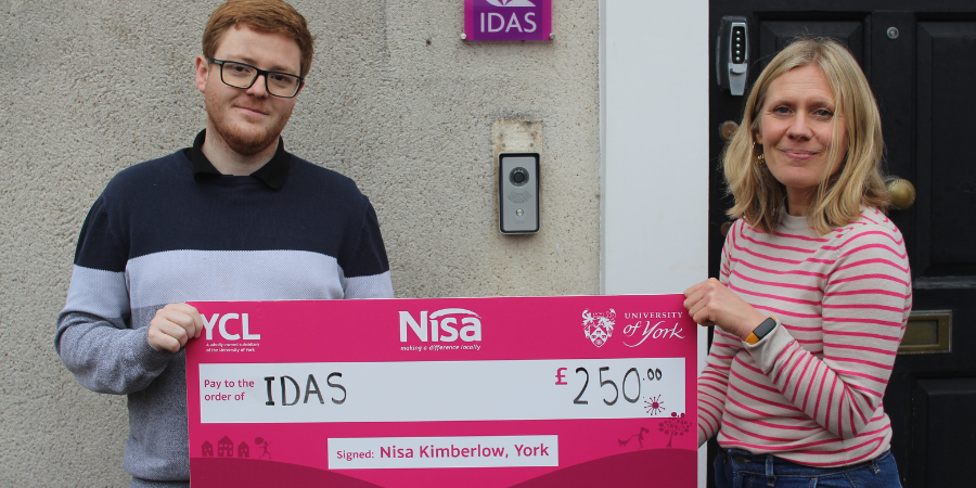 A man from the University of York and a woman from IDAS stand outside an entrance holding a large £250 cheque made out to IDAS, with an IDAS sign on the wall behind them.